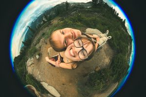 Two friends take a fun fisheye lens selfie outdoors with mountains and green trees in the background.