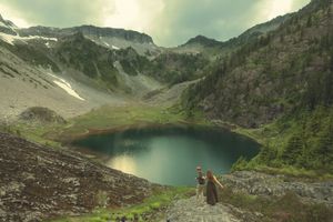 Two hikers stand on a rocky overlook gazing at a stunning alpine lake surrounded by mountains and lush green forests.