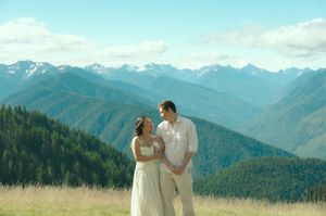 Couple in white attire embracing in a mountain meadow with scenic peaks and green forests in the background.