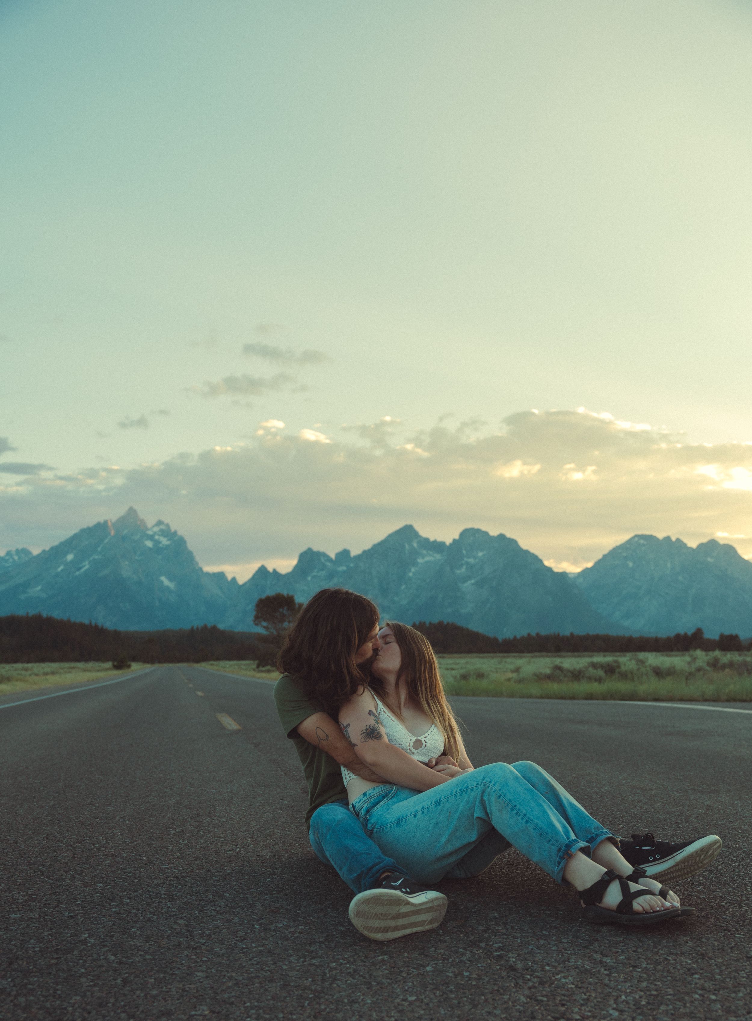 Two people share a tender moment sitting on an empty road with dramatic Grand Teton mountains silhouetted at sunset.