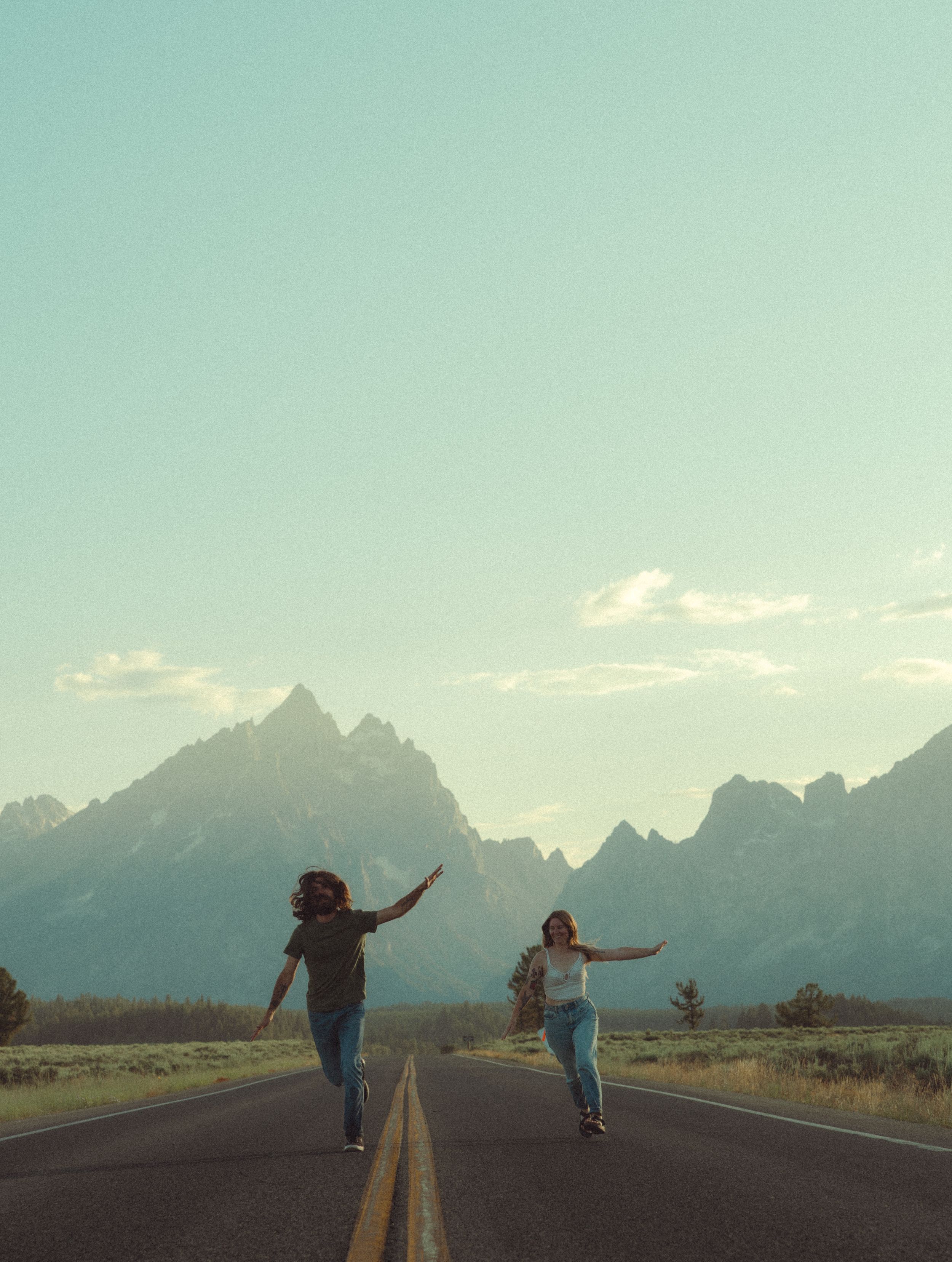 Two people run joyfully down an empty road with the majestic Grand Teton mountains rising in the background under blue skies.
