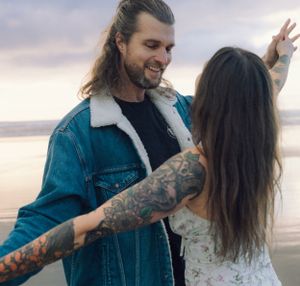 Couple dancing on beach at sunset, man in denim jacket with tattoos.