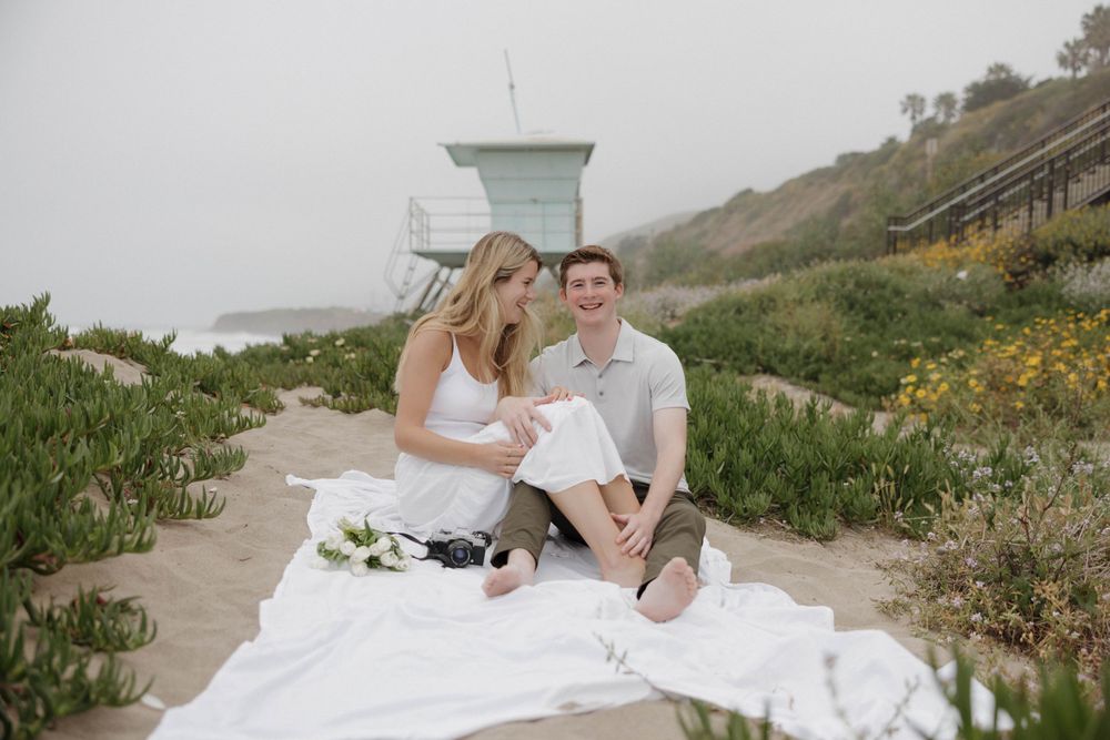 Coastal picnic scene with couple and snacks near turquoise lifeguard tower.