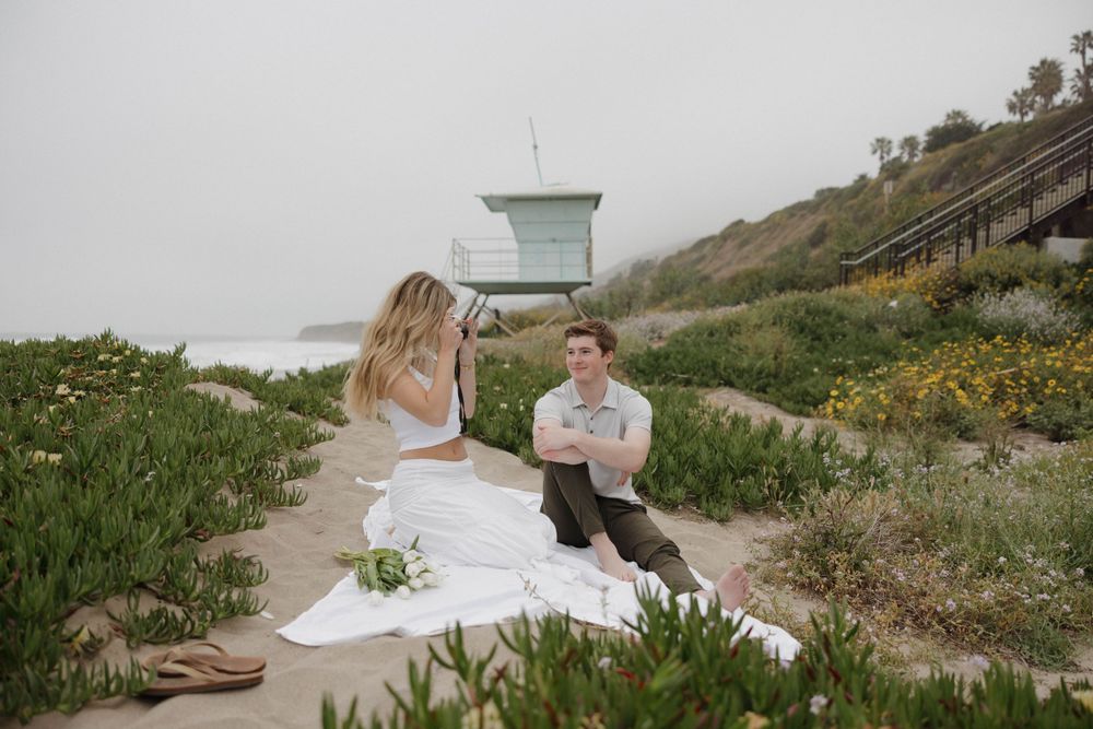 A romantic coastal photoshoot with a couple sitting near a lifeguard tower on a flower-dotted hillside.