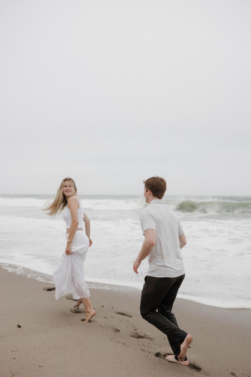 A playful couple walking barefoot along a foggy beach shoreline.