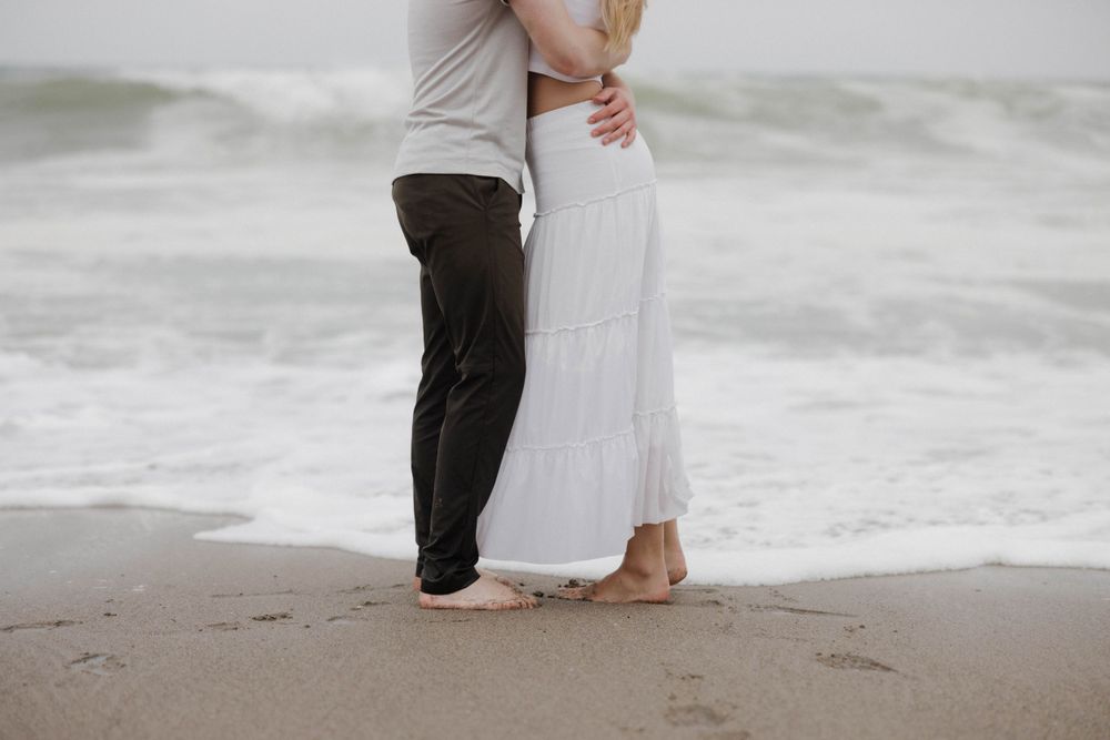 A close-up shot of a couple's legs standing in the sand at the ocean's edge.