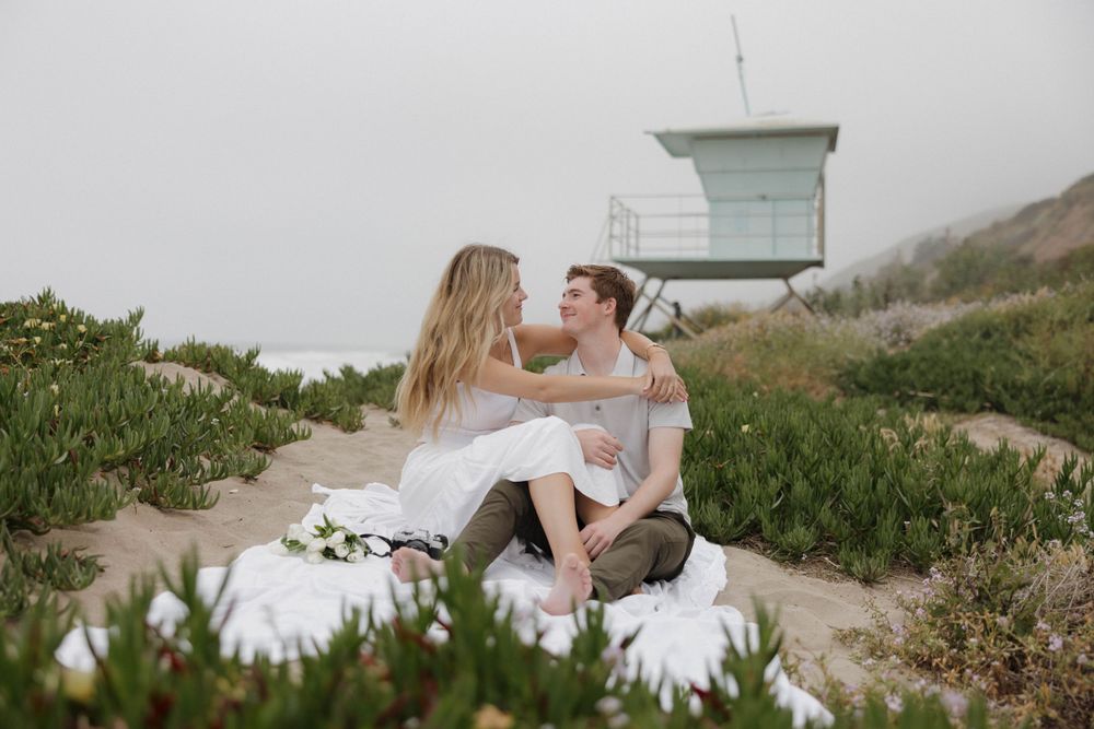 A couple sharing a tender moment near a lifeguard tower on a coastal hillside.
