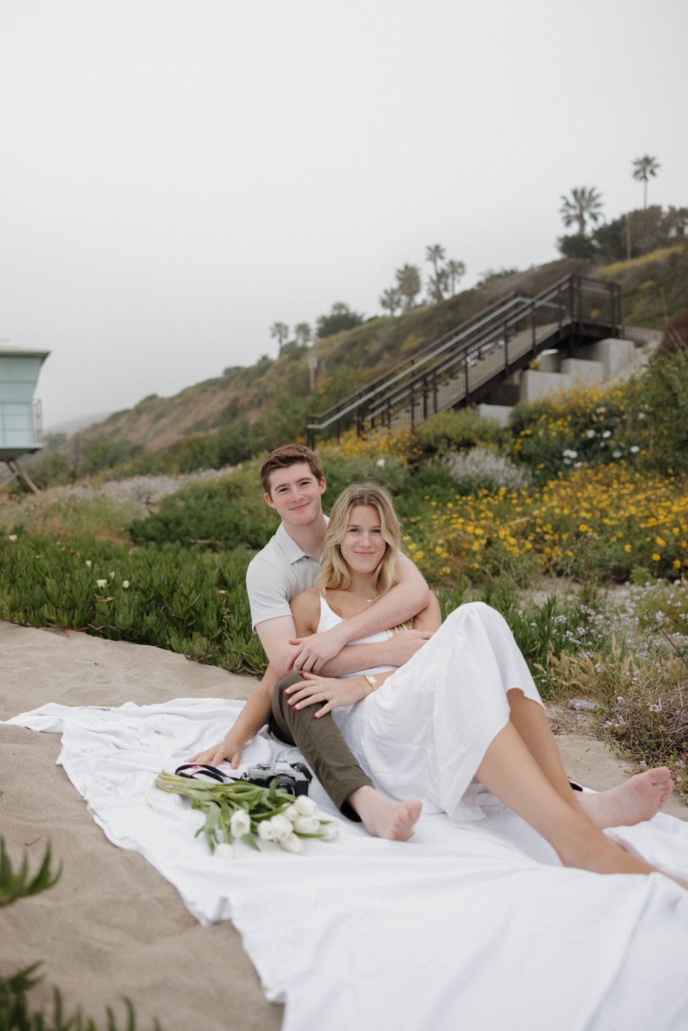 A couple relaxing on a white blanket with coastal flowers and stairs in the background.