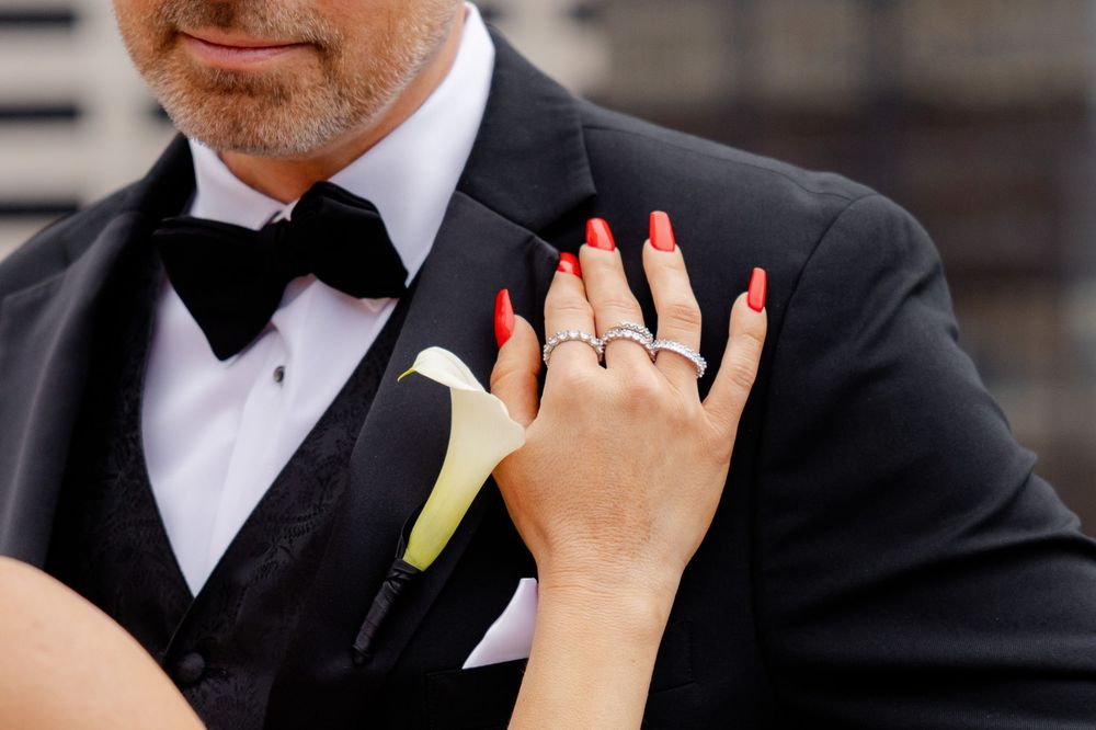 A hand with red nails and rings rests near a black tuxedo with white shirt and bowtie with a calla lily boutonniere.