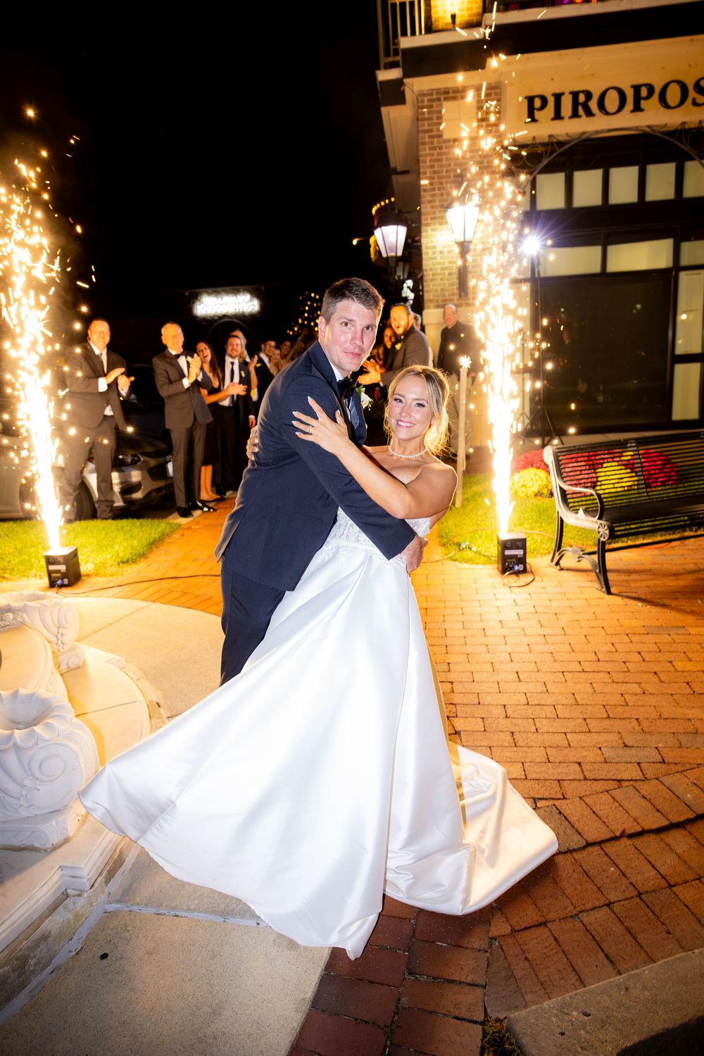 A couple in wedding attire dances together on brick pavement with sparkler fountains illuminating the nighttime celebration.