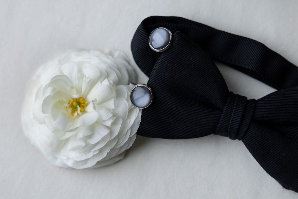 A classic black bow tie decorated with a delicate white rose and pearl buttons against a light gray background.