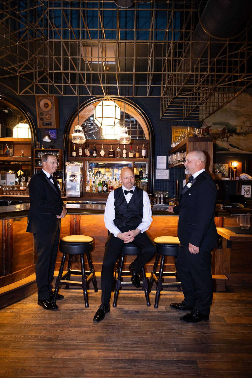 Three people in formal attire stand together at a classic wooden bar with vintage decor and warm lighting.