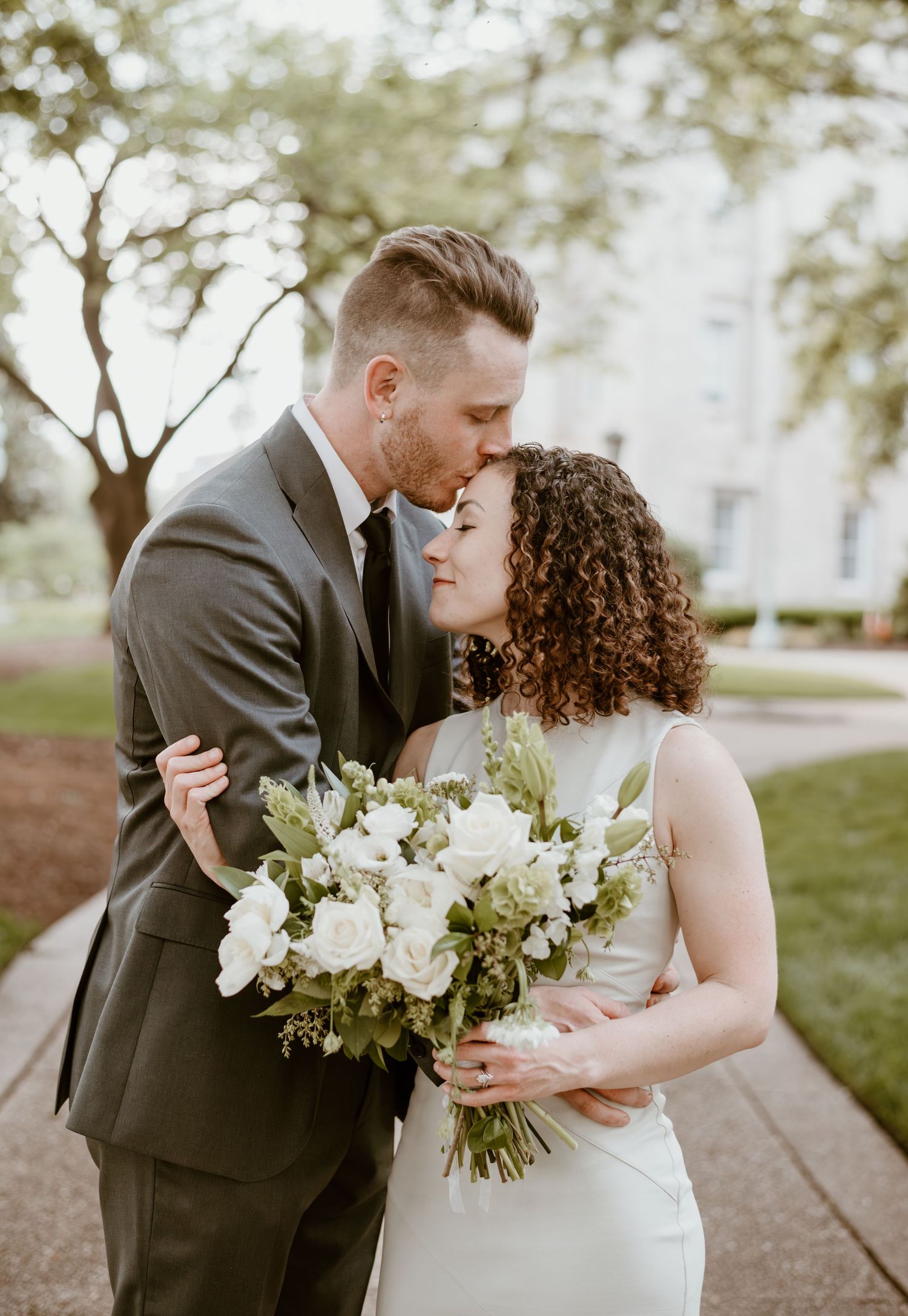 A romantic wedding couple shares tender moments in an outdoor garden setting with the bride holding a white floral bouquet.