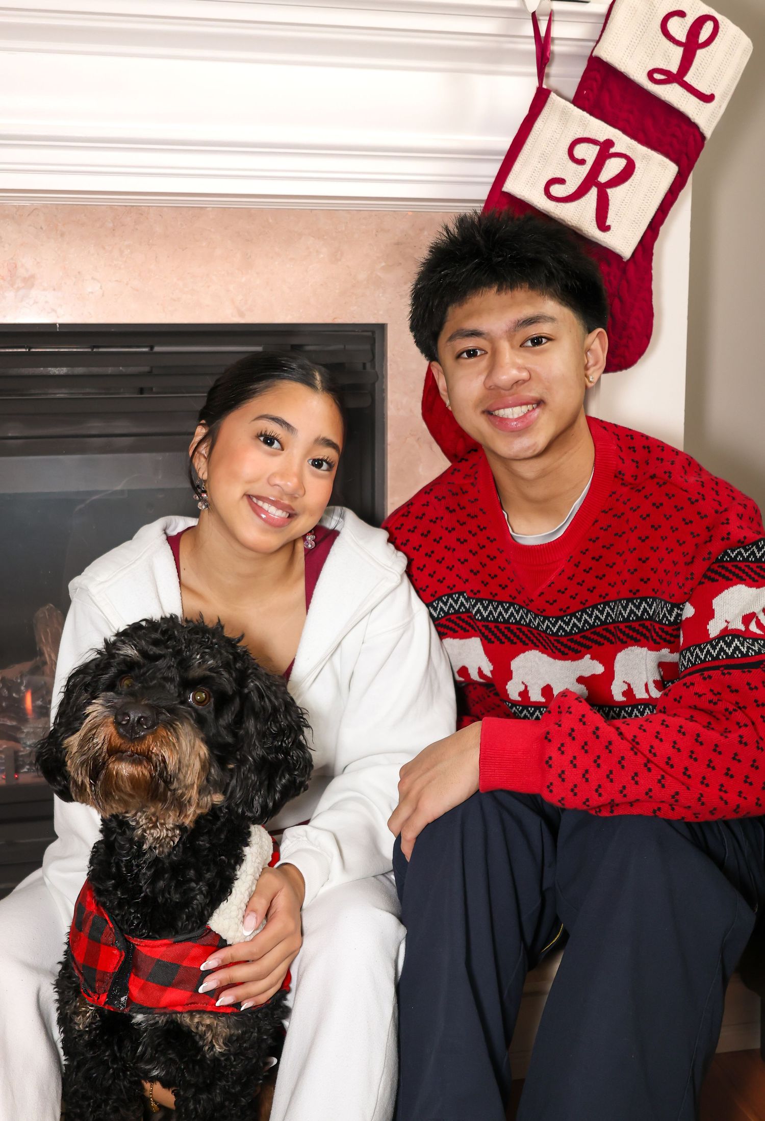 Siblings posing for a Christmas photo next to a fireplace with their family dog