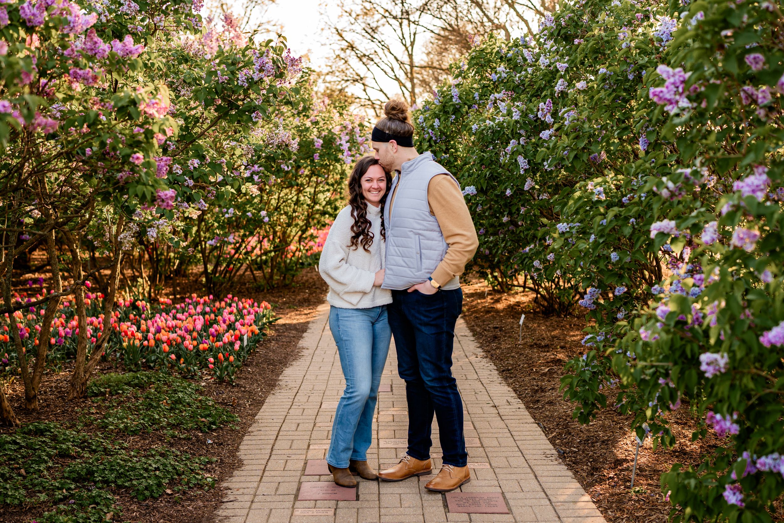 Two people stand close together on a garden path surrounded by blooming pink flowers during golden hour.