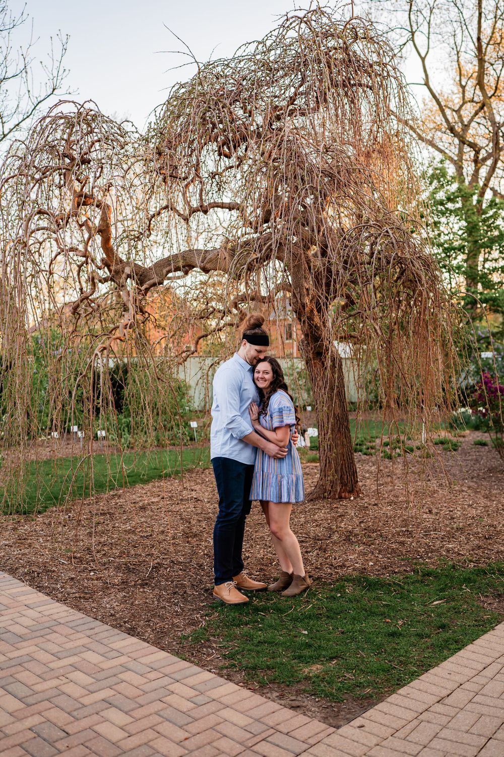 A couple embraces under a weeping willow tree beside a brick pathway in a serene garden setting at dusk.