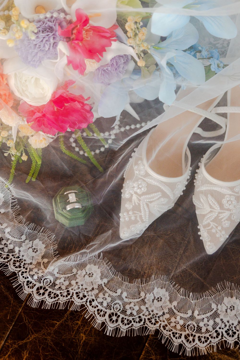 White lace bridal shoes, pearl necklace, ring box, and colorful wedding bouquet arranged on a lace veil.