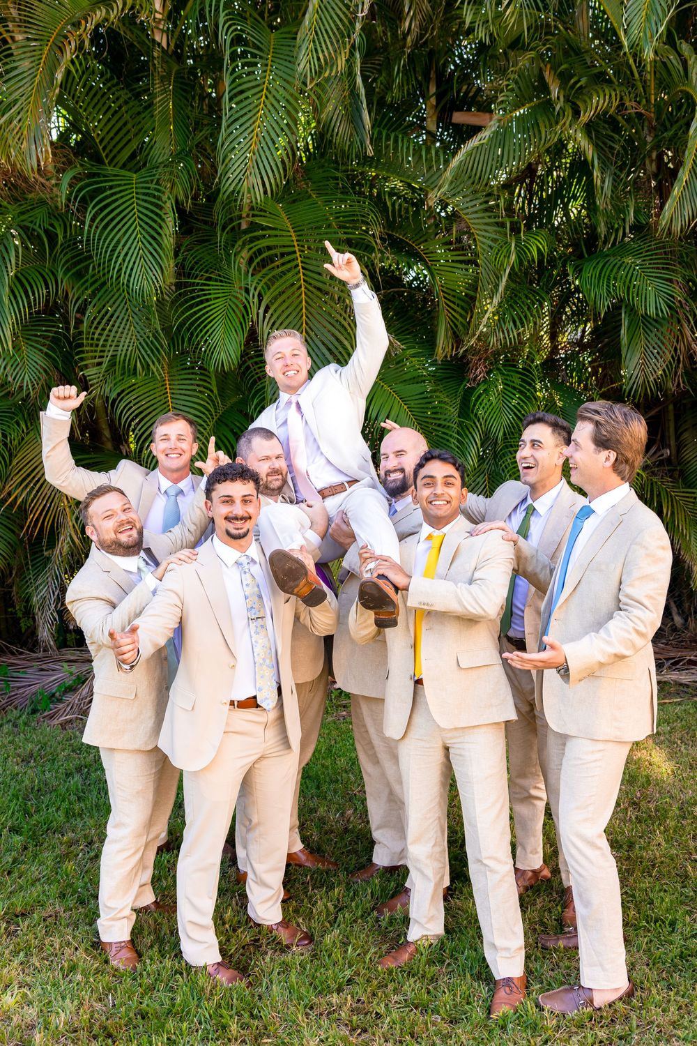 Groomsmen in tan suits lifting groom in celebration outdoors with tropical palm trees in background.