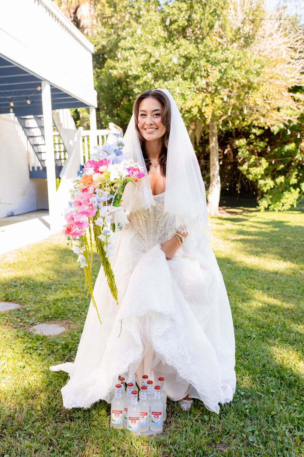 Smiling bride in white gown and veil holds colorful bouquet and fan outdoors, with water bottles at her feet.