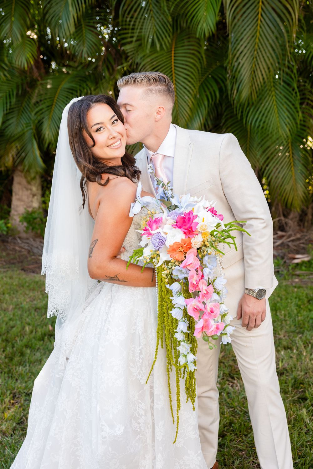 Bride and groom share a kiss outdoors, she holds a colorful cascading bouquet, both dressed in wedding attire near tropical palms.