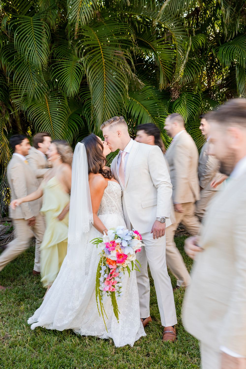 Bride and groom share a kiss surrounded by wedding party in tropical garden setting with lush palm trees.