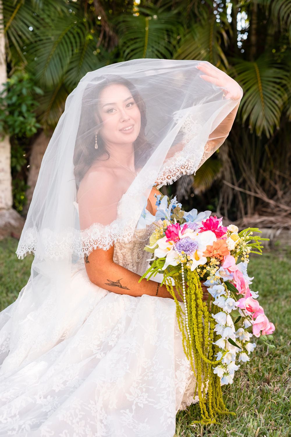 Bride in lace wedding gown holding colorful bouquet with veil lifted, seated outdoors near tropical palm trees.