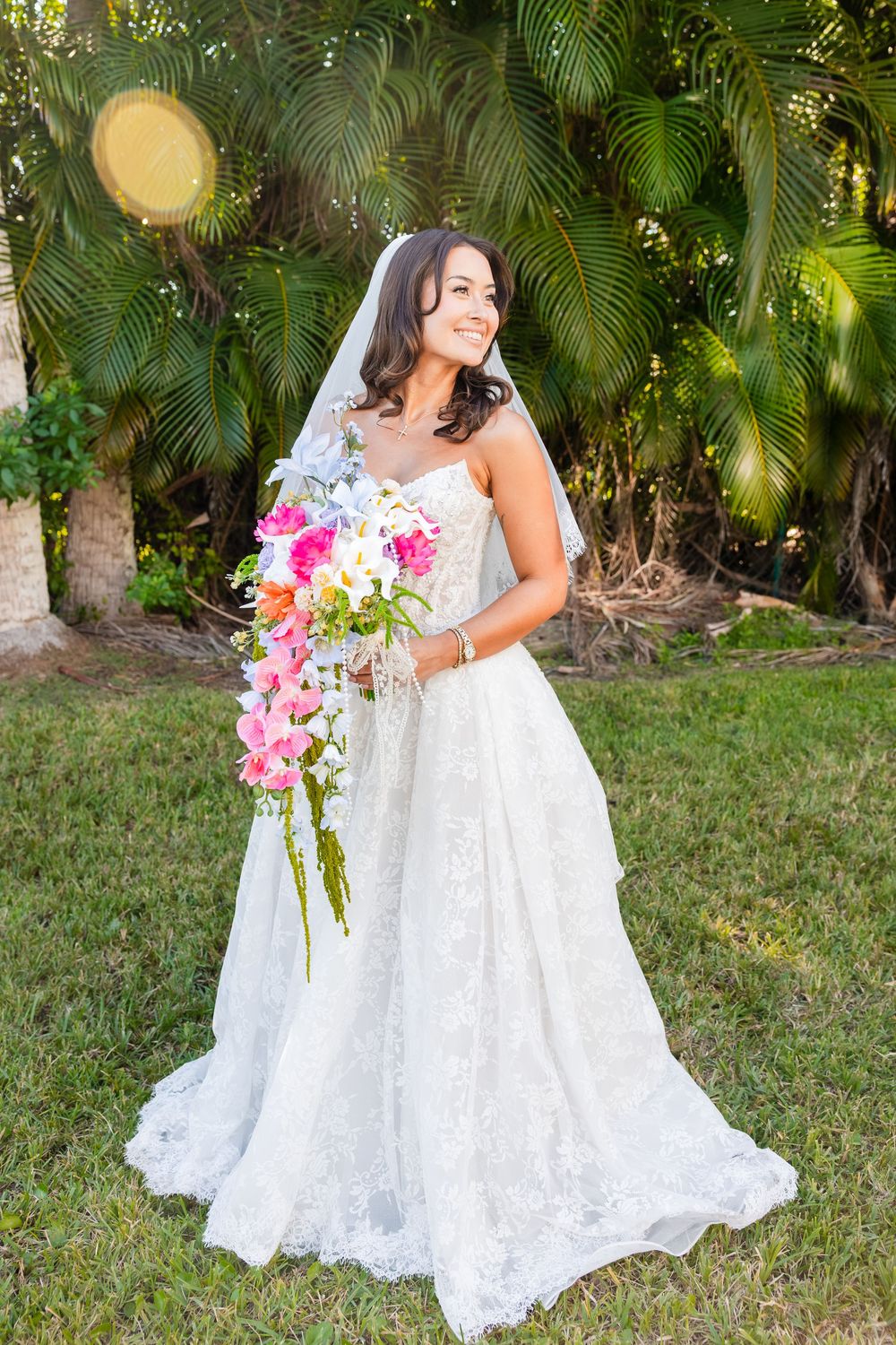 Smiling bride in white lace gown holding colorful cascading bouquet, standing outdoors near tropical palm trees.