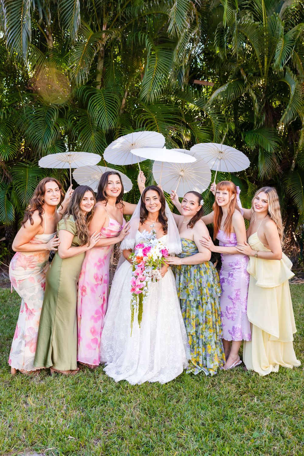 Bride in white gown holds bouquet surrounded by bridesmaids in colorful floral dresses holding white parasols outdoors.