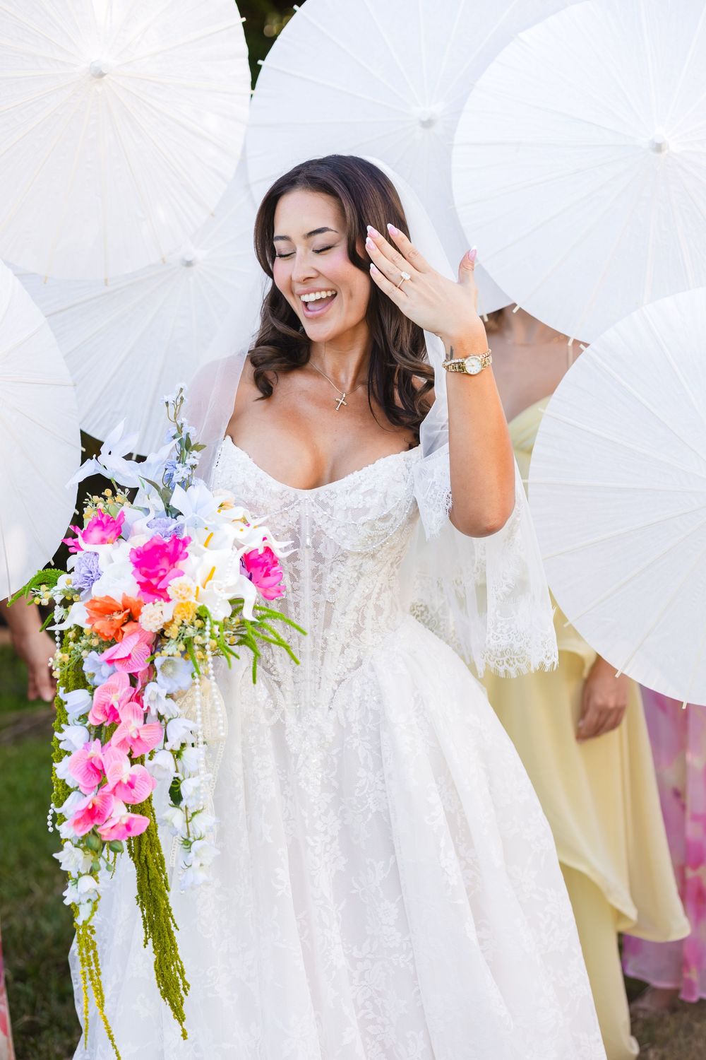 Radiant bride in lace gown holds colorful bouquet, laughing among white parasols at outdoor wedding.