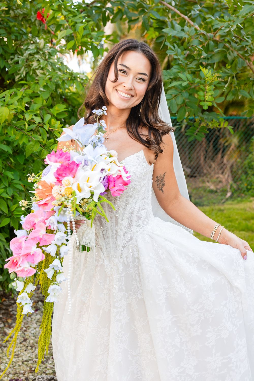 Smiling bride in white lace gown holds colorful cascading bouquet of pink and white flowers outdoors.