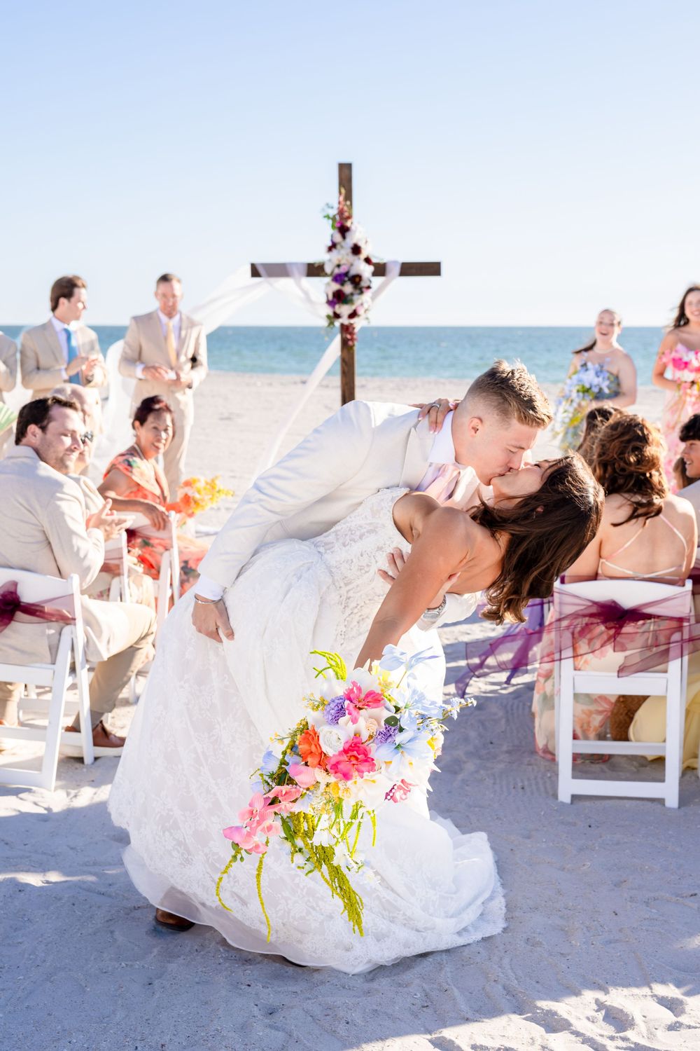 Groom dips bride for romantic kiss at beach wedding ceremony with cross altar and seated guests.