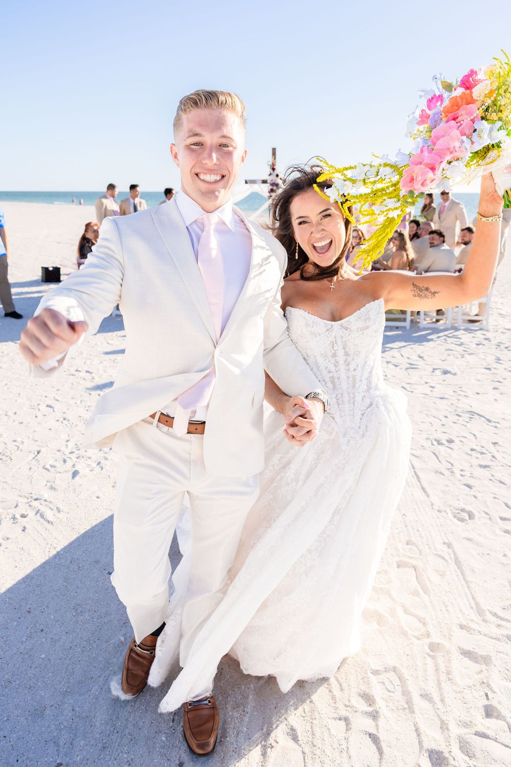 Happy newlyweds celebrate on a sunny beach wedding, bride in white gown holding colorful bouquet, groom in cream suit.