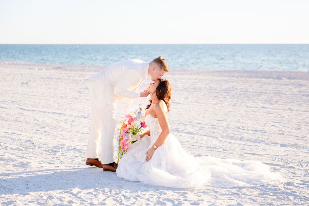 Bride and groom share a romantic kiss on a white sand beach, with the bride holding a colorful bouquet.