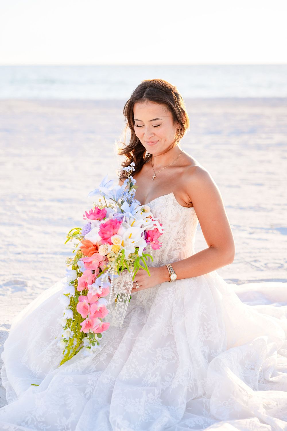 Bride in white lace gown holding colorful cascading bouquet on sandy beach at sunset.