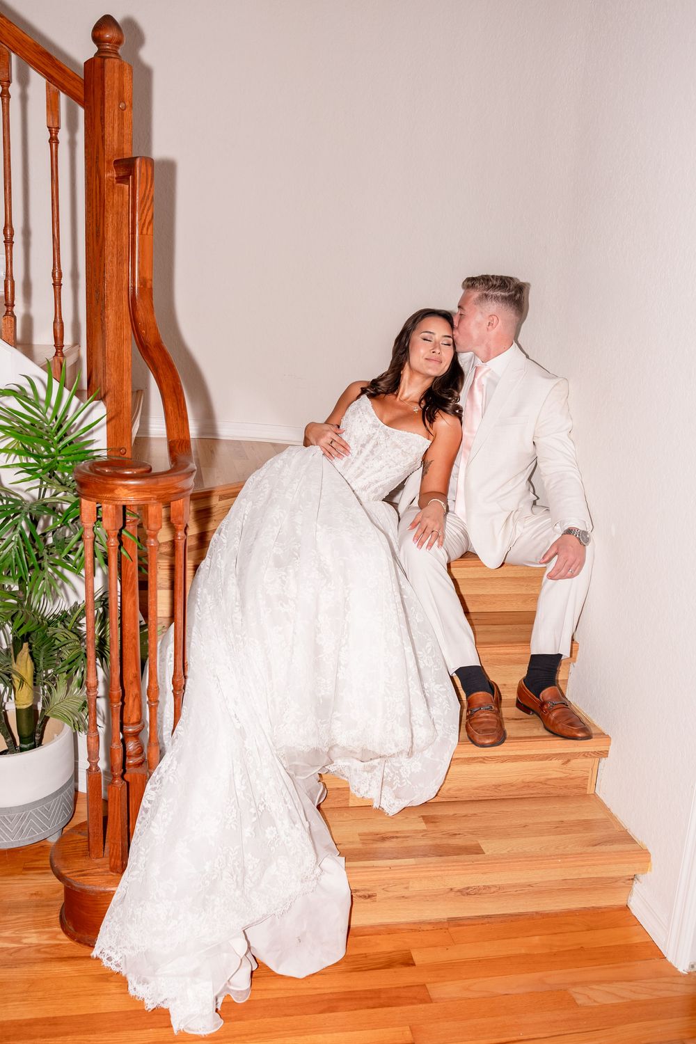 Bride and groom share intimate moment seated together on wooden staircase inside elegant home.