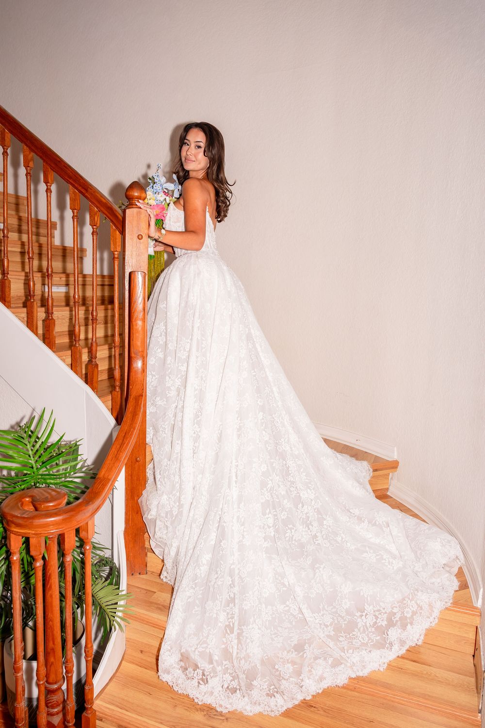 Bride in elegant lace wedding gown with long train holds bouquet on wooden staircase.