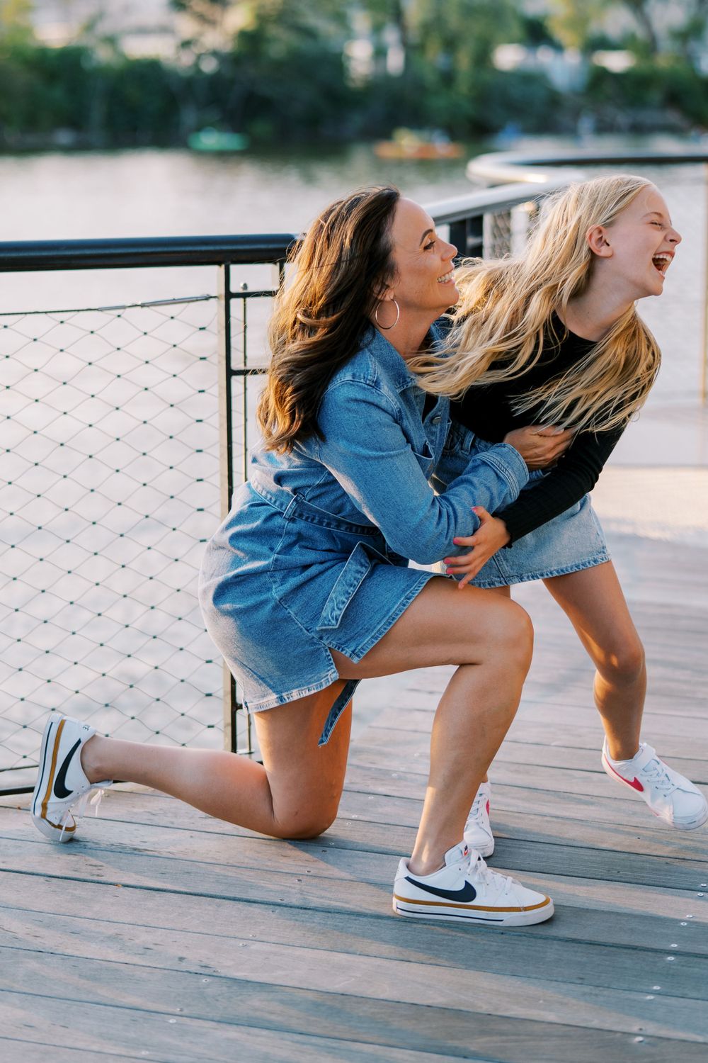 Two people in denim and sneakers crouch playfully on a waterfront boardwalk.