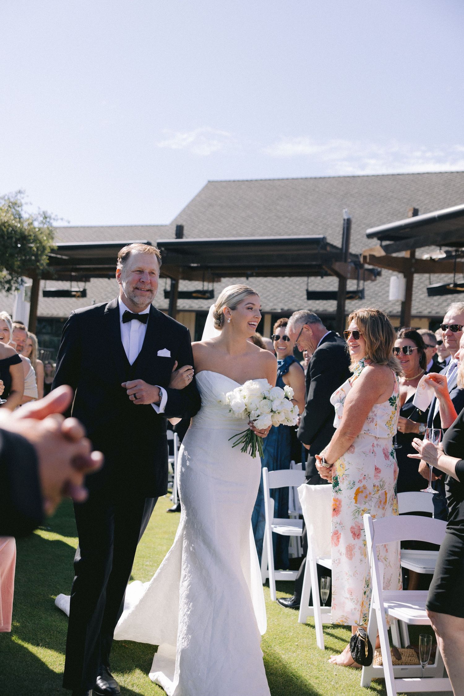 An elegant outdoor wedding ceremony on a sunny day with guests seated on white chairs on a green lawn.