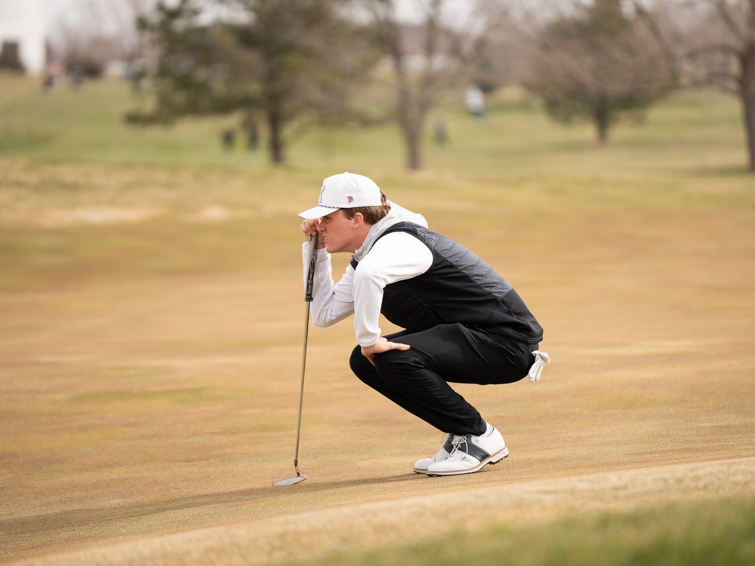 Golfer in white and black attire analyzing putt on brown winter course.