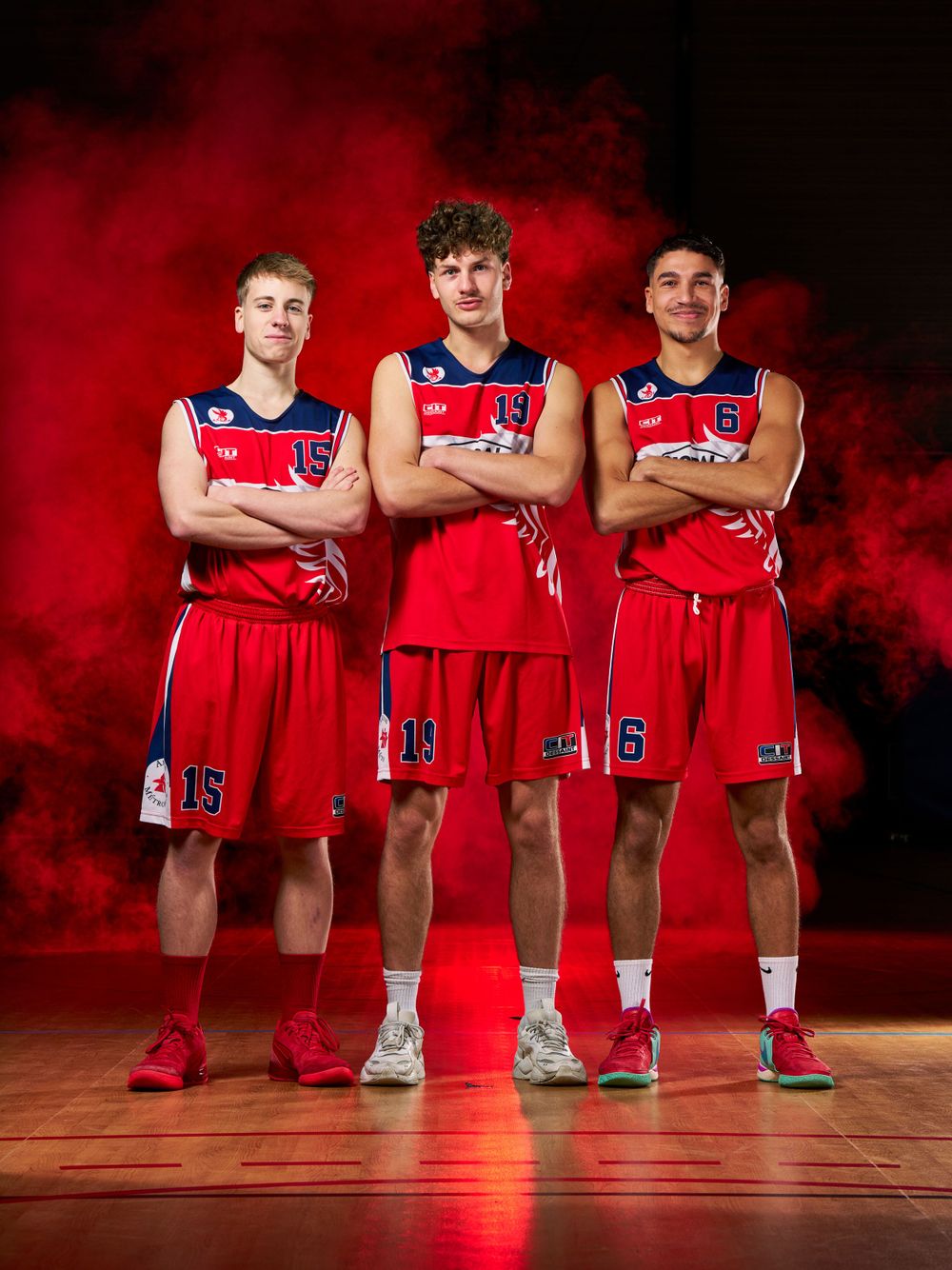 Trois joueurs de basket-ball en uniforme rouge et bleu posent ensemble sur un fond rouge enfumé.