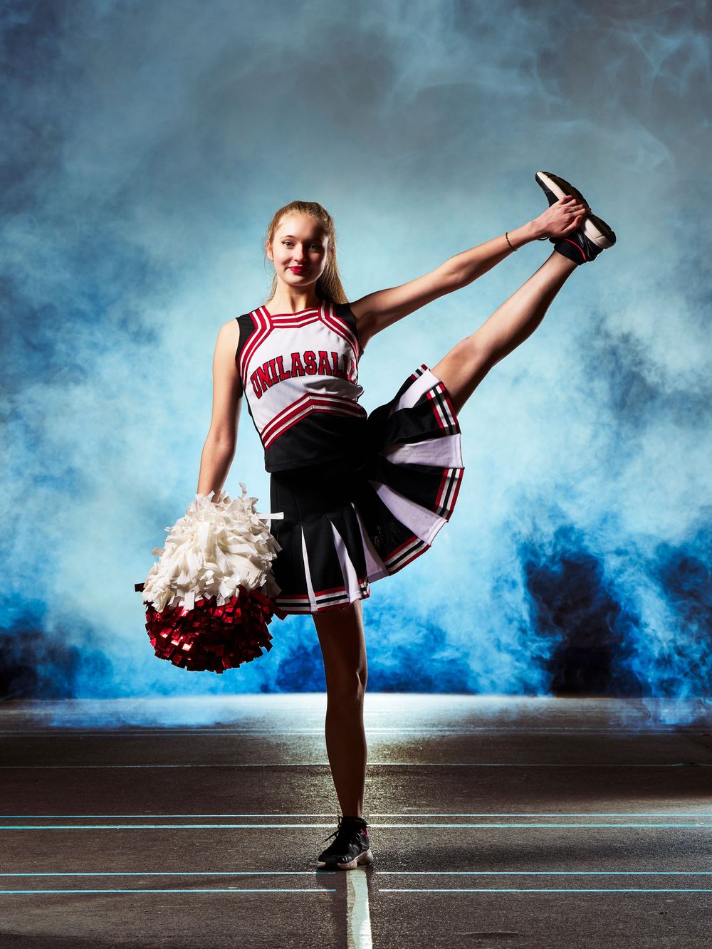 Une pom-pom girl exécute un high kick avec un pom pom sur un fond bleu fumé.