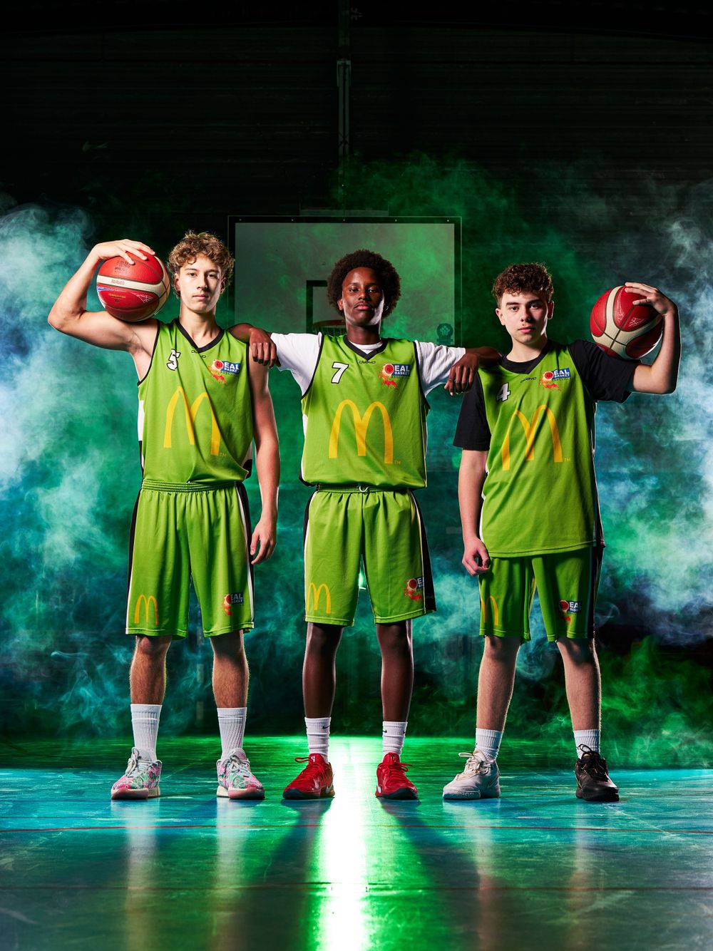 Trois joueurs de basket-ball en uniforme vert vif posent avec des ballons de basket-ball sur un fond vert fumé.