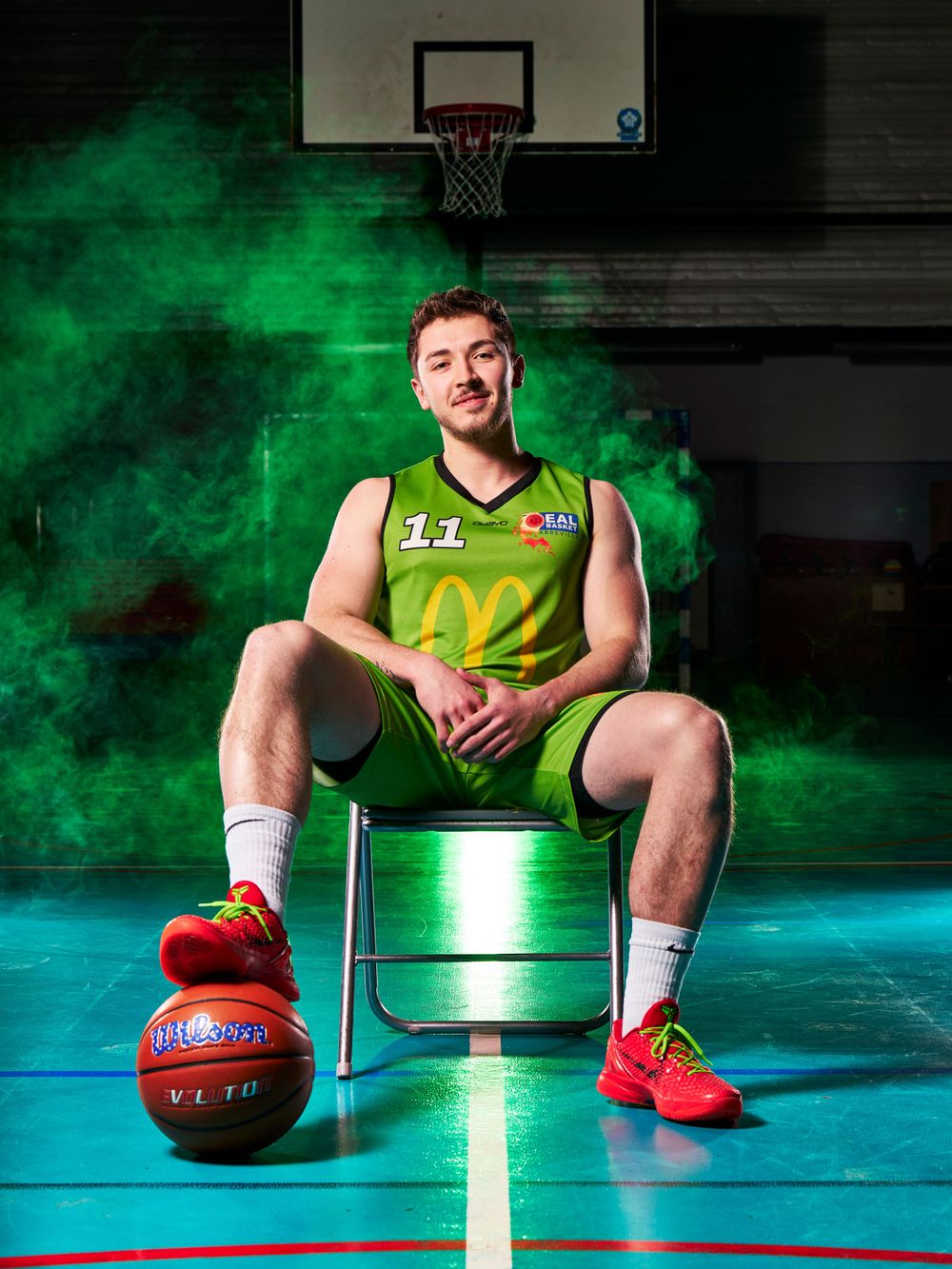 Un joueur de basket-ball en uniforme vert est assis sur une chaise avec un ballon sur fond de fumée verte.