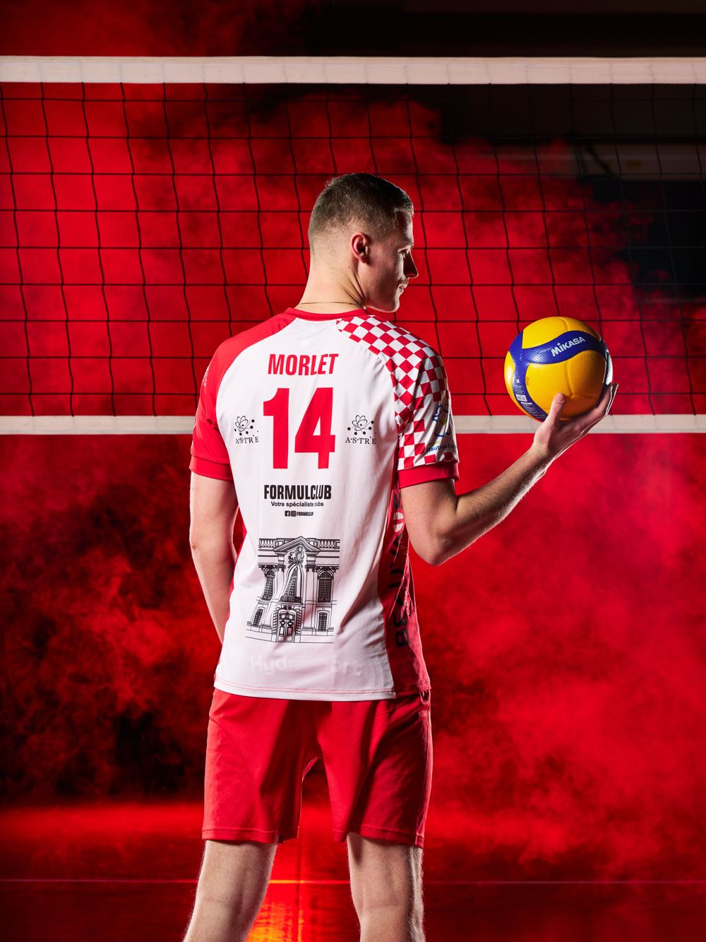 L'athlète en maillot rouge numéro 14 tient le volley-ball devant le filet avec un éclairage rouge atmosphérique.
