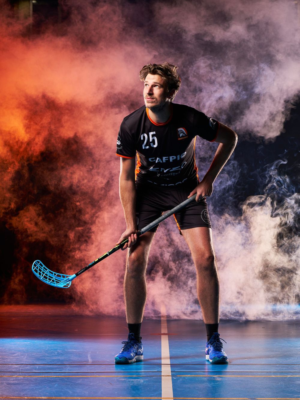 Un joueur de floorball en uniforme noir pose avec un bâton sur un fond orange fumé.
