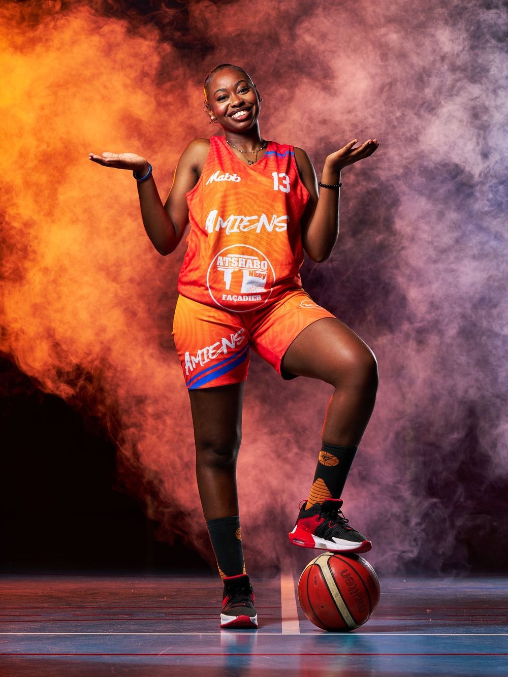 Joueur de basket-ball en uniforme orange pose de manière ludique avec le ballon sur un fond de fumée orange et violet spectaculaire.