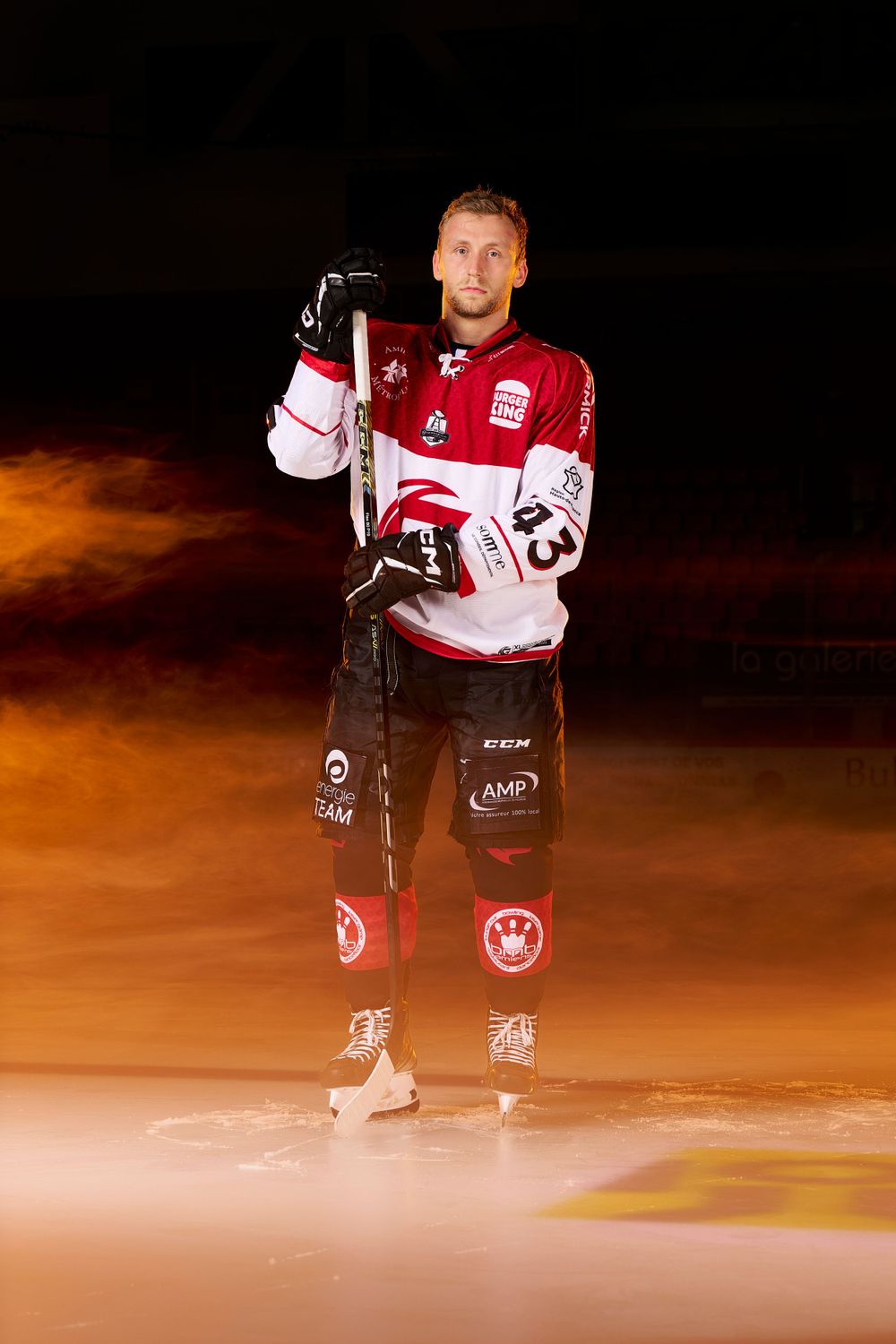 Un joueur de hockey sur glace en tenue rouge et blanche pose sur la glace avec des effets de lumière orange.