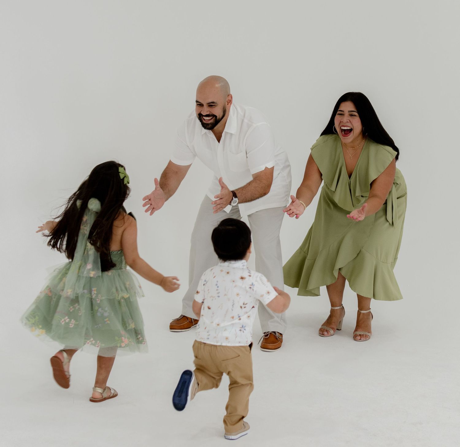 A joyful family of four in white and green outfits playing together in a bright white studio setting.