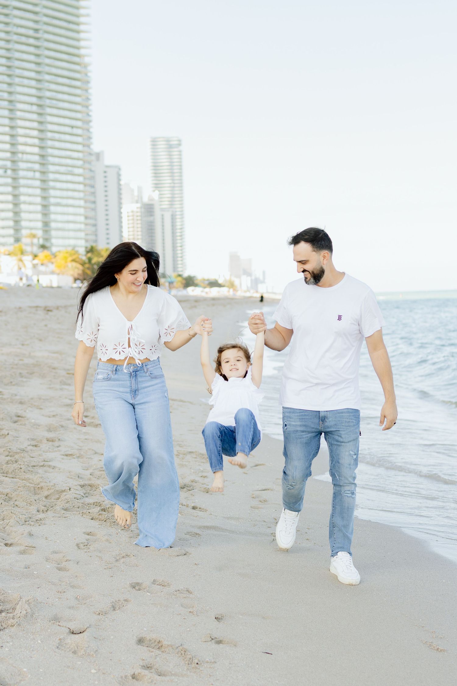 A happy family swings their young child on a sunny beach with city skyline in the background.