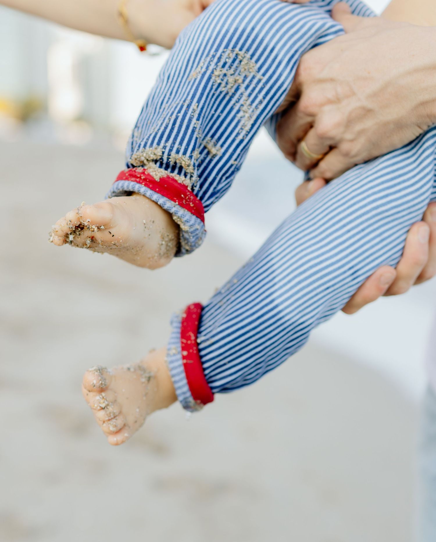 Sandy baby feet in blue striped pants with red trim, held by adult hands at the beach.